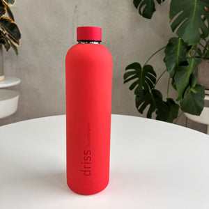 Porter Green driss 1L bottle in Sakai, a red matte body with a red cap and metal neck band, standing on a white table in front of monstera leaves