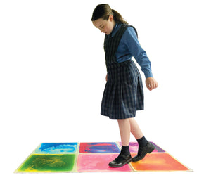 Six square sensory liquid floor tiles laid out on a pale floor with a child standing on one, the coloured liquid spreading out from under the foot.