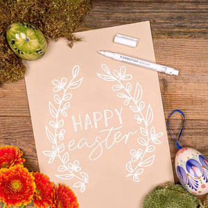 Kraft paper sheet hand lettered HAPPY easter with a white leafy wreath, a Staedtler white pen, decorated eggs and orange flowers on wood