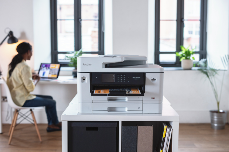 Brother office printer on a shared workspace shelf with printed documents in a bright modern office.