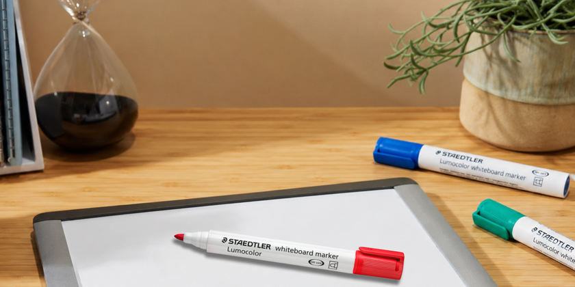 Staedtler Lumocolor whiteboard markers arranged on a desk with a small whiteboard, marker caps, stationery and plant for everyday board writing.