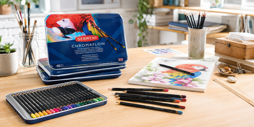 Derwent Chromaflow coloured pencils and tins arranged on a creative studio desk with artwork, sketchbook, brushes and drawing supplies