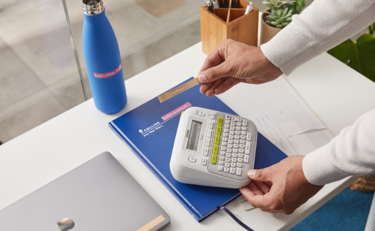 Label maker being used to organise folders, drink bottles and desk items in a clean office workspace.
