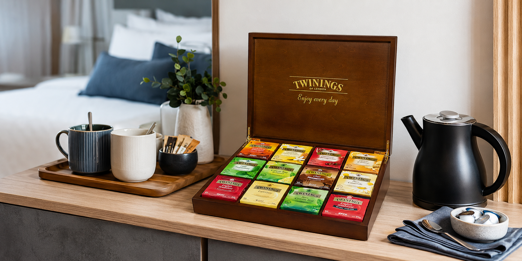 Premium guest tea amenities box with assorted tea sachets, mugs and kettle in a modern hotel room refreshment area