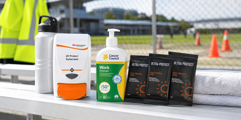 Workplace sunscreen bottles, sachets and dispenser arranged at an outdoor safety station near a sports field.