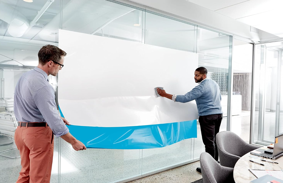 Self-adhesive dry erase whiteboard film being applied to a glass office wall.