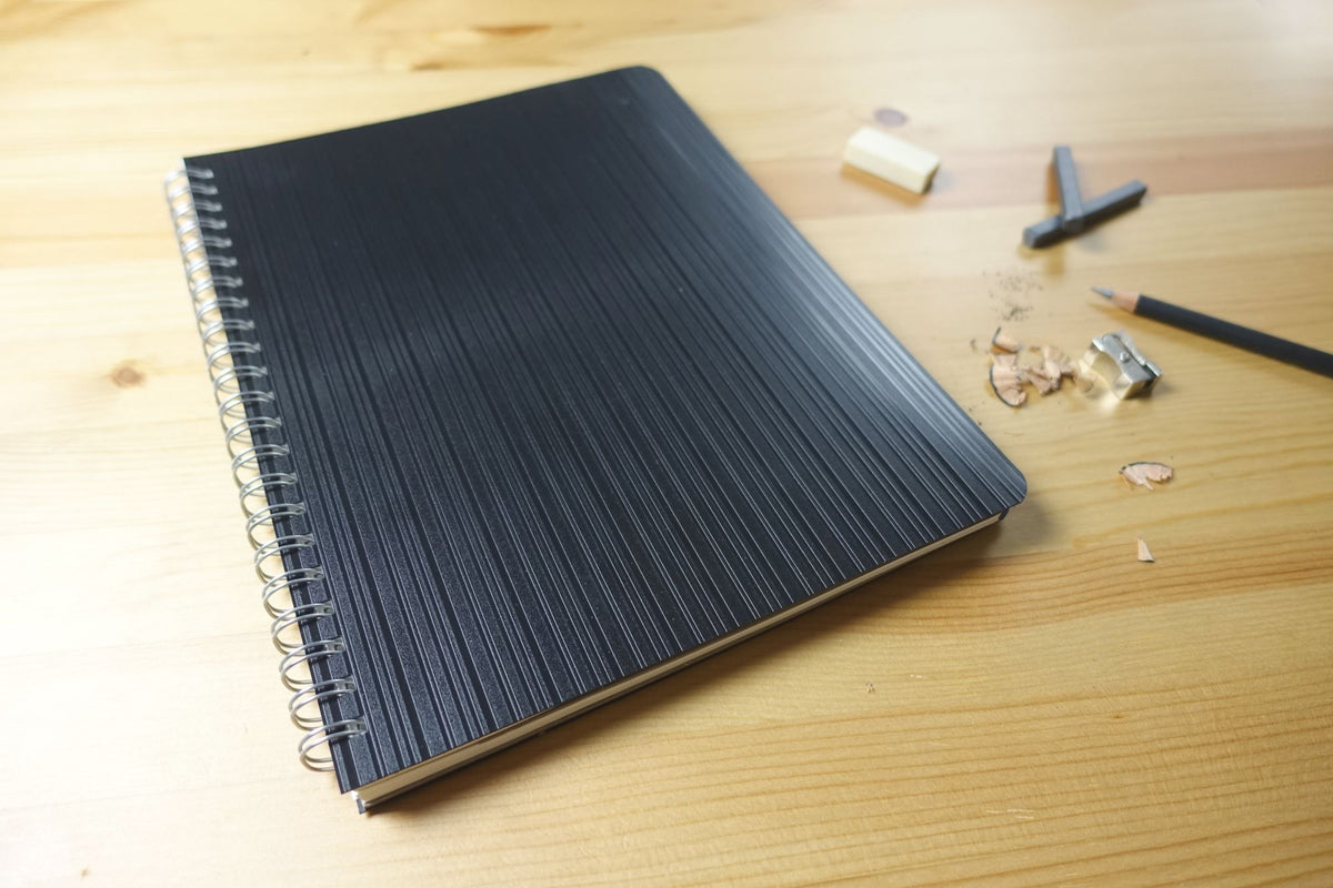 Quill Premium Visual Art Diary closed on a pine desk, showing the black striped cover and spiral binding beside a pencil, an eraser, a sharpener and pencil shavings