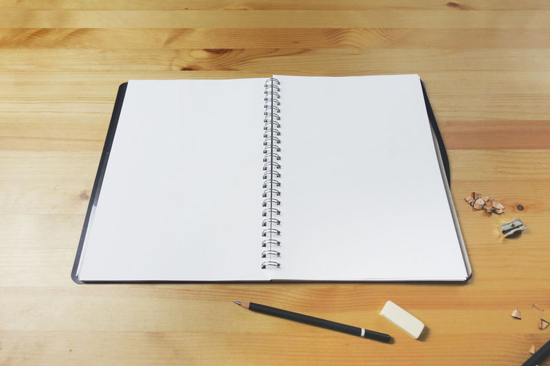 Quill Premium Visual Art Diary lying open on a pine desk at a blank double page spread, black covers either side of the central spiral, with a pencil and an eraser in front