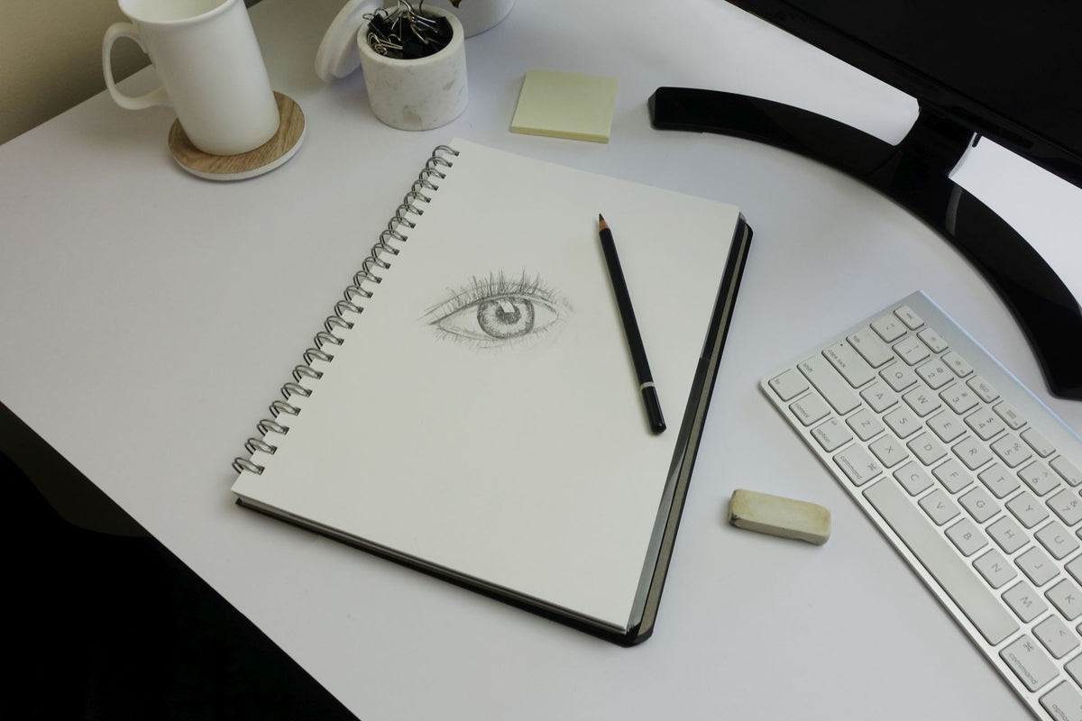 Quill Premium Visual Art Diary lying open on a white desk with a pencil drawing of an eye on the page, next to a keyboard, a mug, sticky notes and an eraser