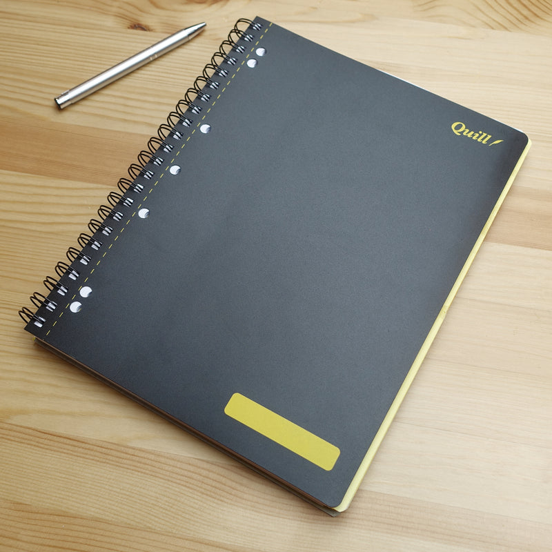Quill Q Series 3 subject book closed on a wooden desk with a pen beside it