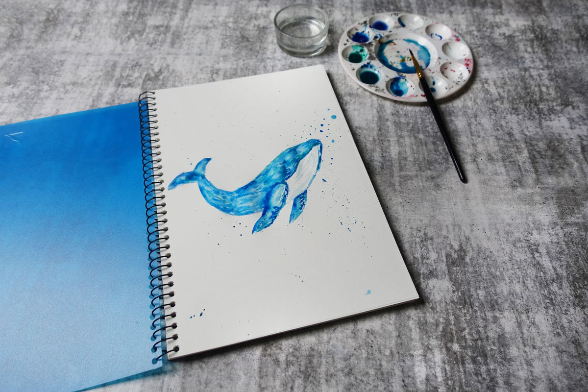 Quill Visual Art Diary A4 open on a grey surface showing a watercolour whale painted on the white cartridge page, with a paint palette and brush beside it