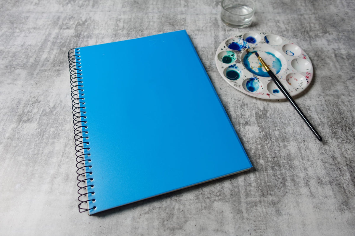 Closed Quill Visual Art Diary A4 with a Marine Blue cover lying on a grey surface beside a round paint palette and a brush