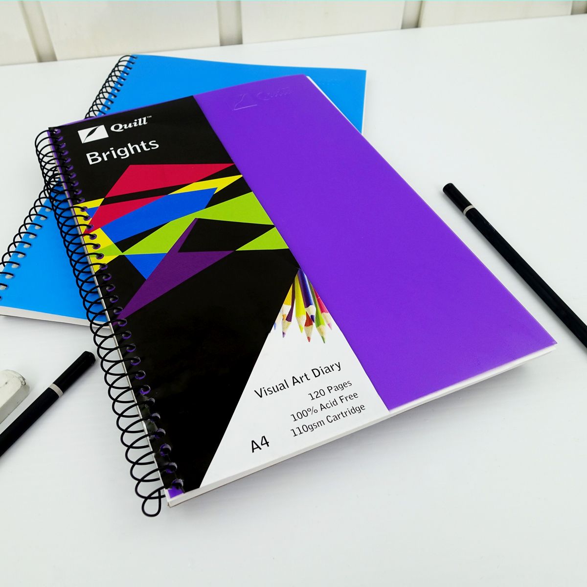 Quill Visual Art Diary A4 Dark Purple with its printed band, resting on a second diary on a white desk with pencils and an eraser