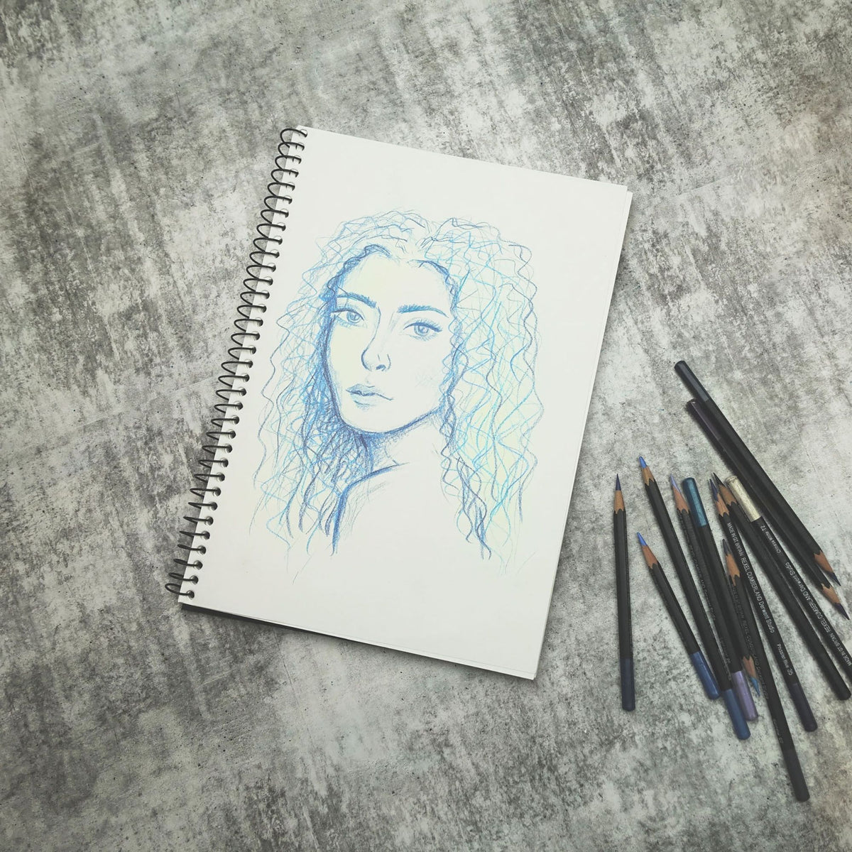 Quill Visual Art Diary A4 open to a blue and green pencil portrait drawing, with a bundle of coloured pencils on a grey surface