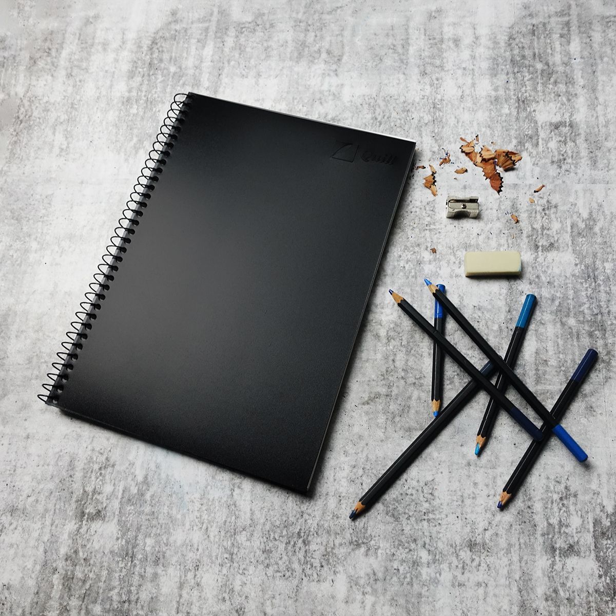 Closed Quill Visual Art Diary A4 with a black cover on a grey surface, with pencils, a sharpener and an eraser beside it