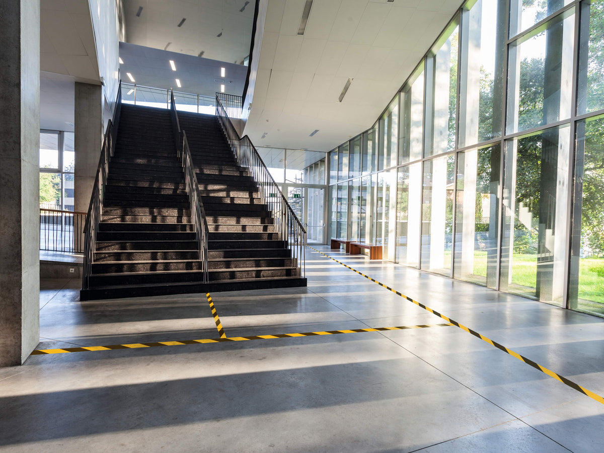 Yellow and black DURALINE 50/07 tape marking the floor area at the foot of a staircase in a glazed foyer