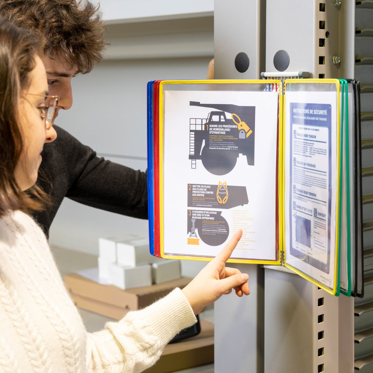 Tarifold A4 wall display mounted on a grey workshop column, a person pointing to an instruction sheet in the yellow pocket while a colleague looks on
