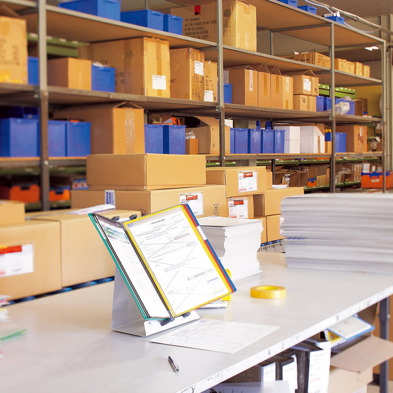 Tarifold A4 desk display stand on a warehouse packing bench, assorted coloured pockets holding a printed form in front of shelving and cartons