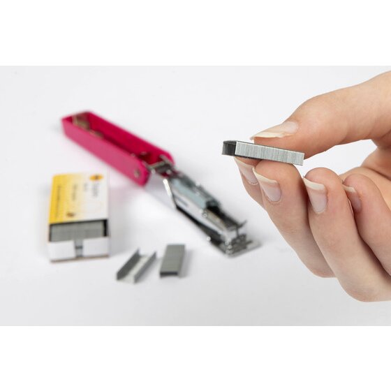 A hand holding a strip of No 10 staples beside an open pink mini stapler, a small yellow staple box and loose staples