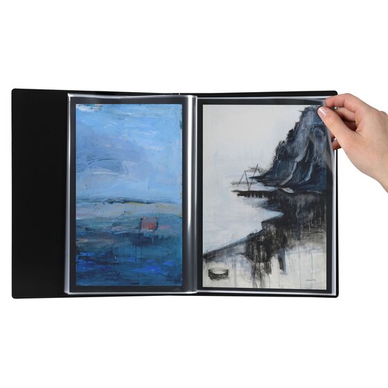 The black Marbig A3 refillable display book opened flat on a white background with two landscape paintings displayed in facing clear A3 pockets and a hand turning the right hand pocket.
