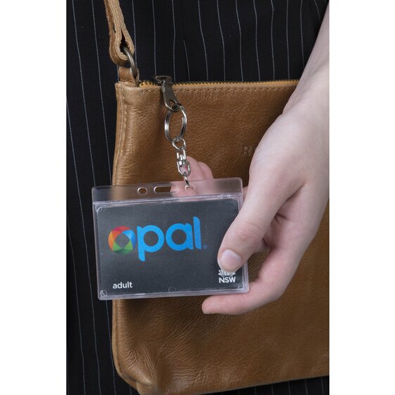 Rexel clear rigid card holder clipped by its chain to a tan shoulder bag strap, with a hand holding a transport card inside