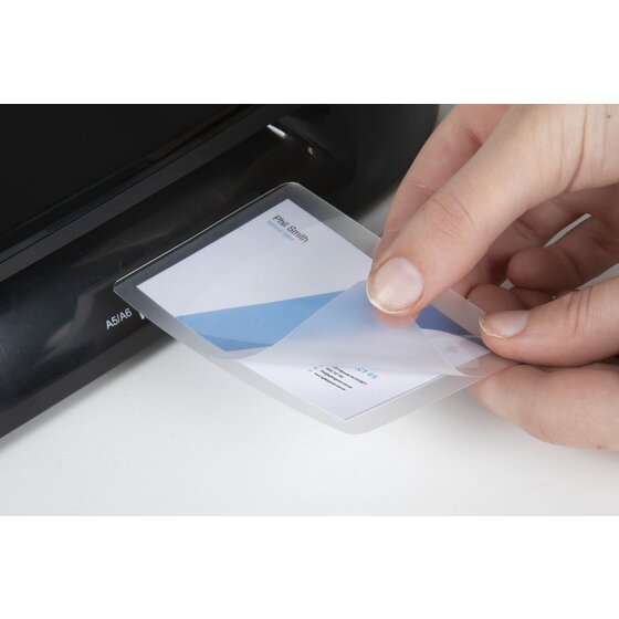 A hand feeding a small laminating pouch holding a printed name card into the slot of a black laminator, with the slot marked A5 and A6.