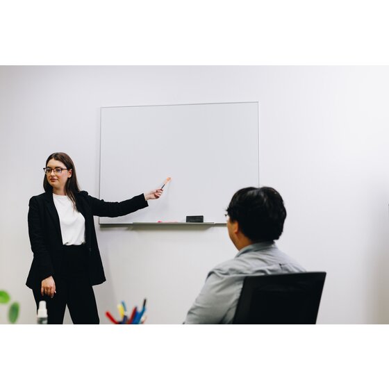 A presenter in a black jacket points to a white wall mounted whiteboard in a silver frame while a colleague in a grey shirt listens, from ACCO's photography for QTPWI1209A.