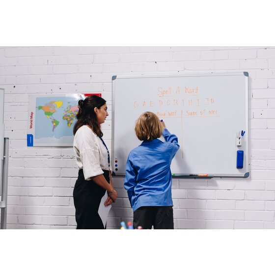 Quartet commercial whiteboard on a white brick wall in a classroom, a woman standing beside a child in a blue shirt who writes orange letters on the board, with a world map poster to the left
