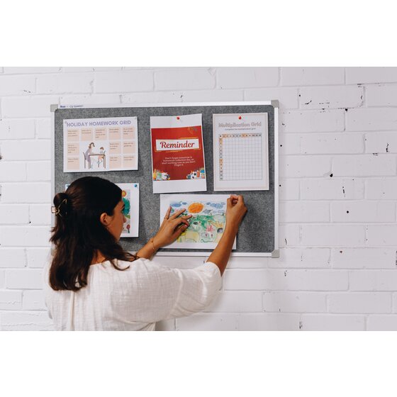 Woman pinning a child's drawing to a grey Quartet Penrite felt pinboard on a white brick wall, beside a homework grid, a reminder notice and a multiplication chart