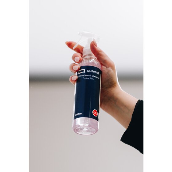 Quartet QTTWC250 Whiteboard Cleaner in use, a hand in a black sleeve holding the navy label 250ml trigger spray bottle against a pale grey background