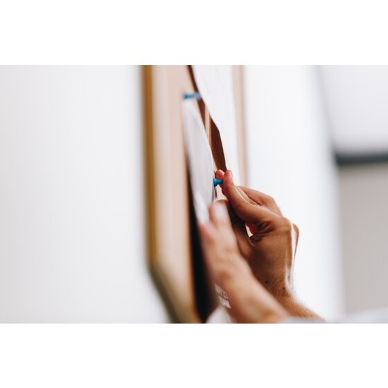 Hand pressing a blue push pin into a sheet on a Quartet pine framed corkboard, photographed close and side on against a white wall