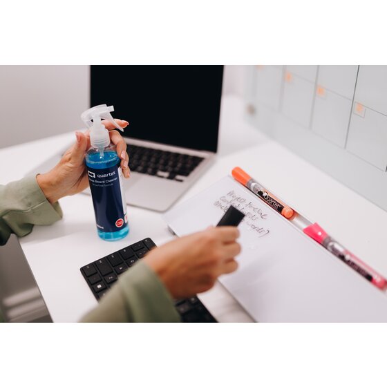 Quartet Glass Board Cleaner QTGC250ML1 in use, a hand holding the navy label spray bottle while another hand wipes a white board surface on a desk beside a keyboard and markers