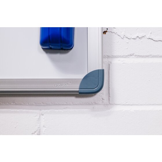 Close up of a Quartet whiteboard corner showing a grey corner cap on an aluminium rail printed with the Quartet name, and a blue eraser on the board against a white brick wall
