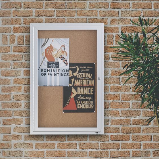 Quartet enclosed cork board in an aluminium cabinet on a brick wall beside a potted palm, with an exhibition poster and a dance festival poster pinned behind the door