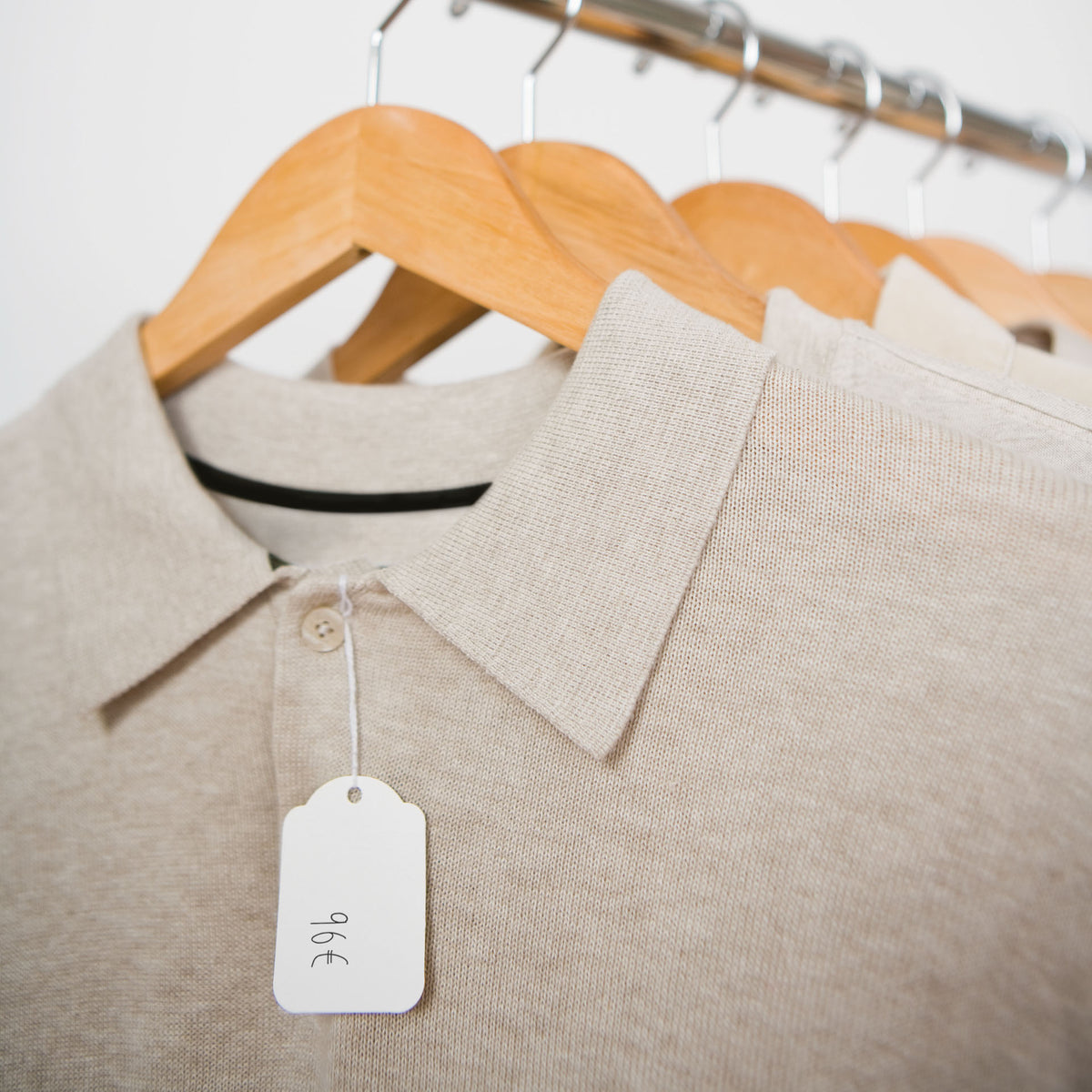 Apli photo of a white strung ticket with a domed top and a handwritten euro price, tied by a thin white cord to the button of a beige knitted polo shirt on a wooden hanger