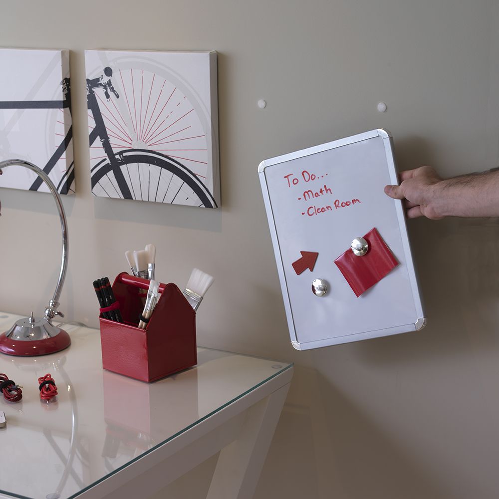 A hand fitting a small whiteboard to a beige wall where two white Velcro Brand circles are stuck, above a glass desk with a red brush caddy and a lamp