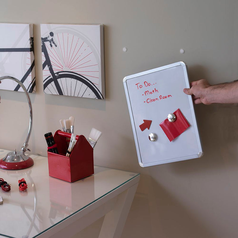A hand fitting a small whiteboard to a beige wall where two white Velcro Brand circles are stuck, above a glass desk with a red brush caddy and a lamp