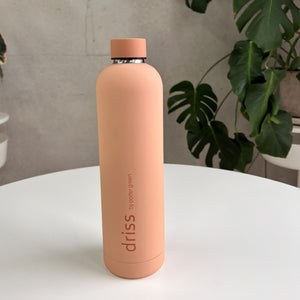 Porter Green driss 1L bottle in Luanda, a soft peach matte body with a deeper peach cap and metal neck band, on a white table with monstera leaves behind