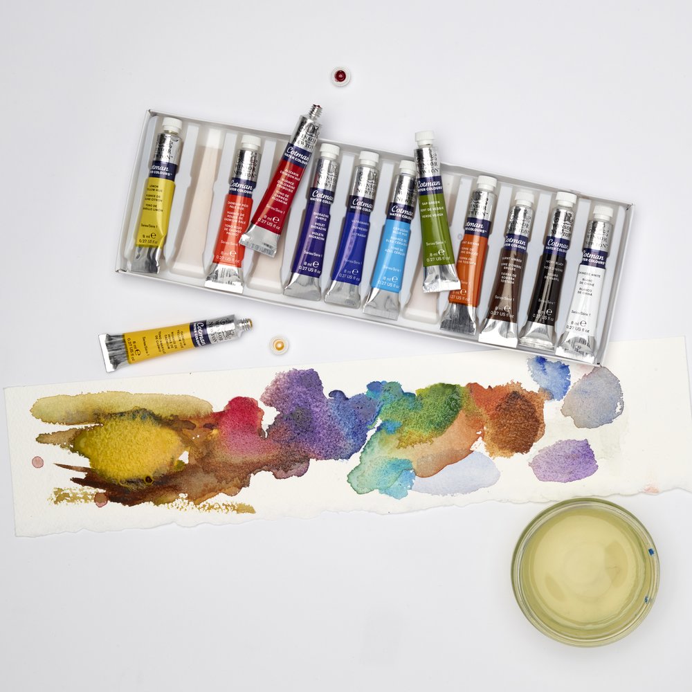 Cotman watercolour tubes from the 12 tube collection beside a strip of paper painted with blended yellow, red, purple, blue, green and brown washes and a water jar