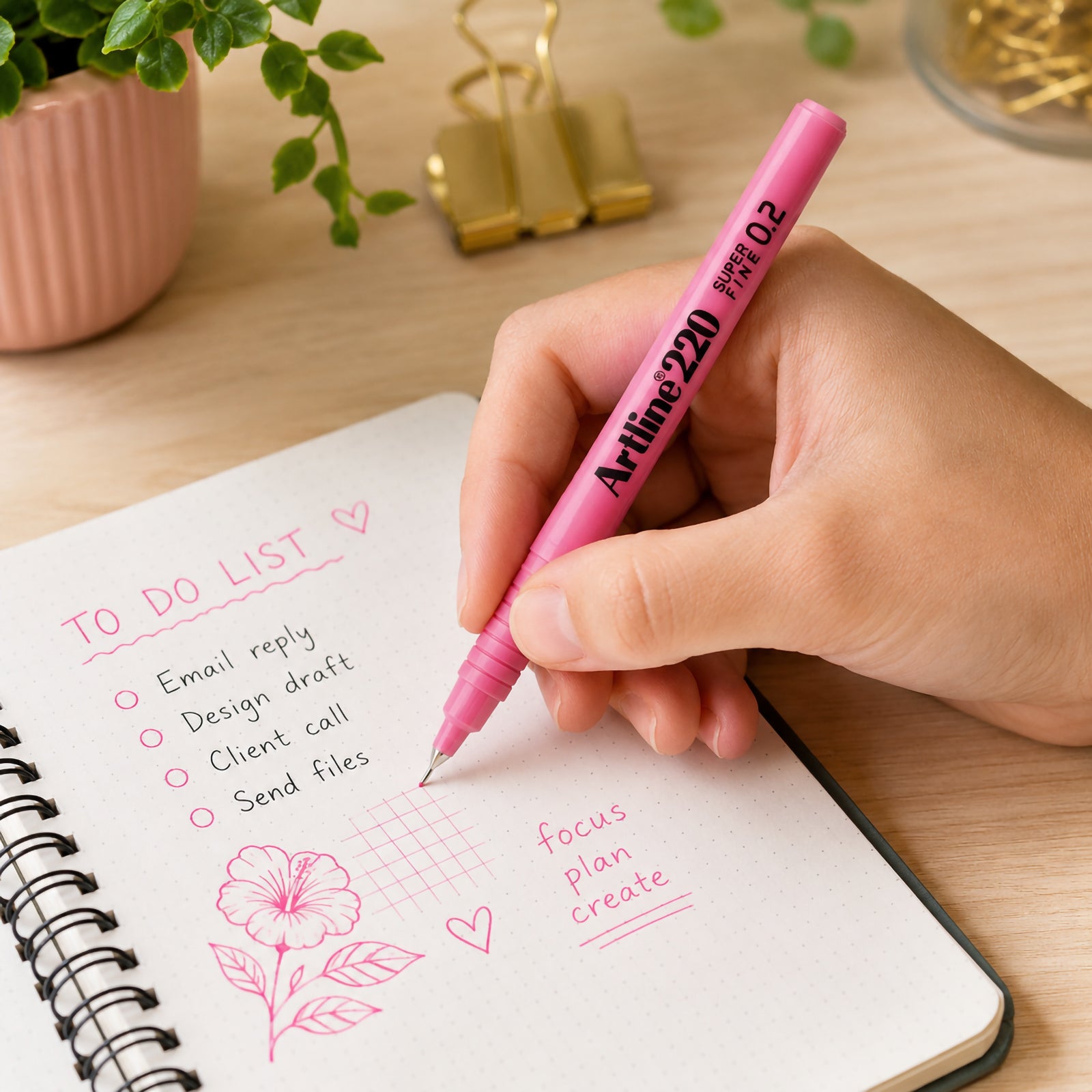 A lifestyle workspace showing the Artline 220 Fineliner Pen 0.2mm Pink being used to write a to do list and create delicate sketches inside a dotted notebook. The ultra fine pink pen demonstrates precise line work on a clean wooden desk with stationery accessories and indoor plants, highlighting its suitability for journalling, planning and creative note taking.