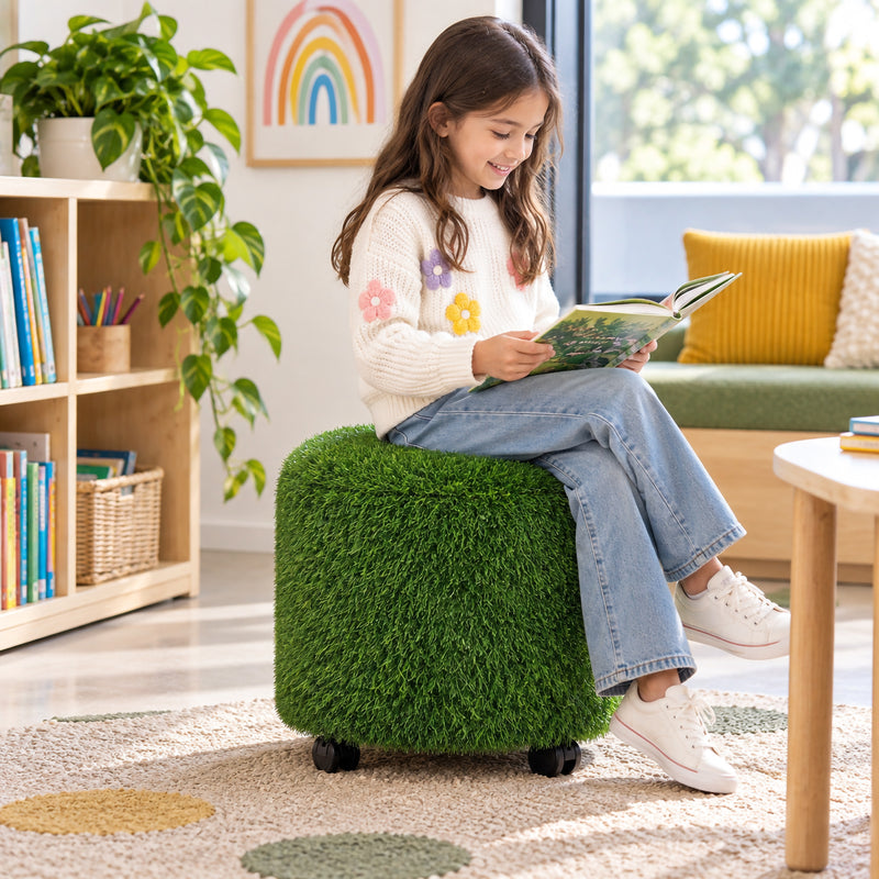 MTA Essentials Aalto Round Grass Ottoman shown in use by a child reading in a bright classroom library setting. The compact green sensory seat features dense artificial grass textured upholstery and black castors beneath the base, creating an inviting flexible seating option for reading corners, sensory areas and relaxed learning spaces.