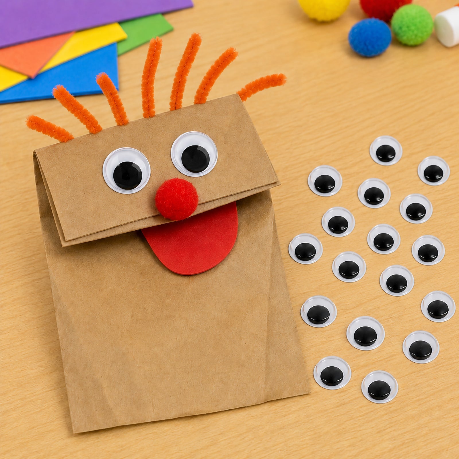 Handmade paper bag puppet featuring adhesive black and white joggle eyes, a red pom pom nose and pipe cleaner hair. The surrounding loose joggle eyes highlight the ease of creating expressive puppet faces for storytelling, classroom projects and imaginative play.