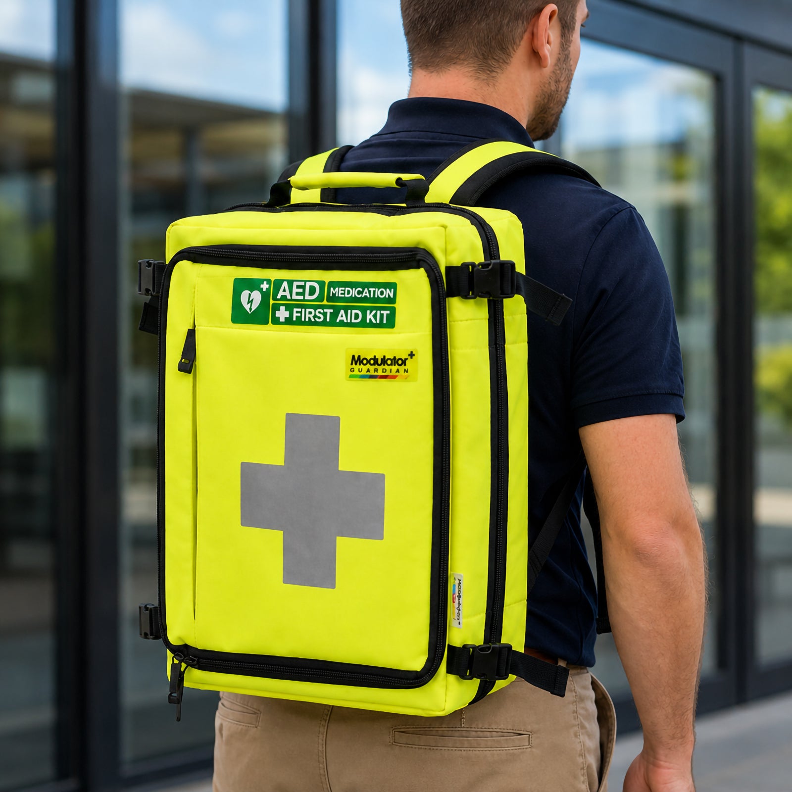 Person wearing the high visibility AeroKit Modulator Guardian First Aid Kit Backpack outside a commercial building, showing the large reflective first aid cross, AED and medication identification labels, shoulder straps and portable emergency response design.