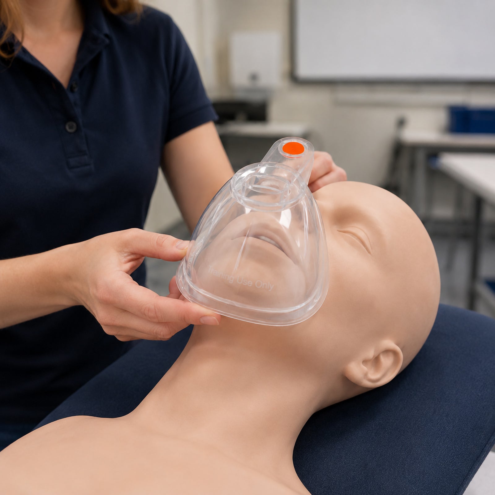 Instructor positioning a clear AeroLearn Adult Training Mask over the nose and mouth of a life sized adult training manikin. The transparent mask has a prominent orange training identifier and demonstrates realistic placement during supervised spacer and inhaler education in a classroom or simulation environment.