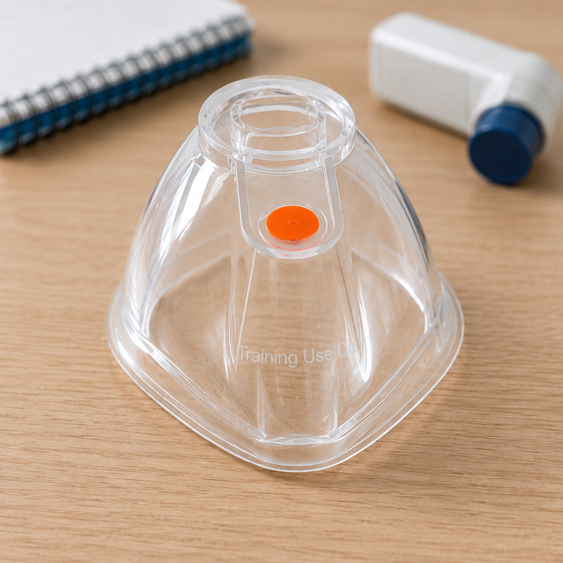 AeroLearn Adult Training Mask displayed upright on a training desk with a notebook and inhaler style training device visible in the background. The transparent adult sized mask features a circular connector opening, orange training indicator and visible Training Use Only marking for straightforward identification during education sessions.