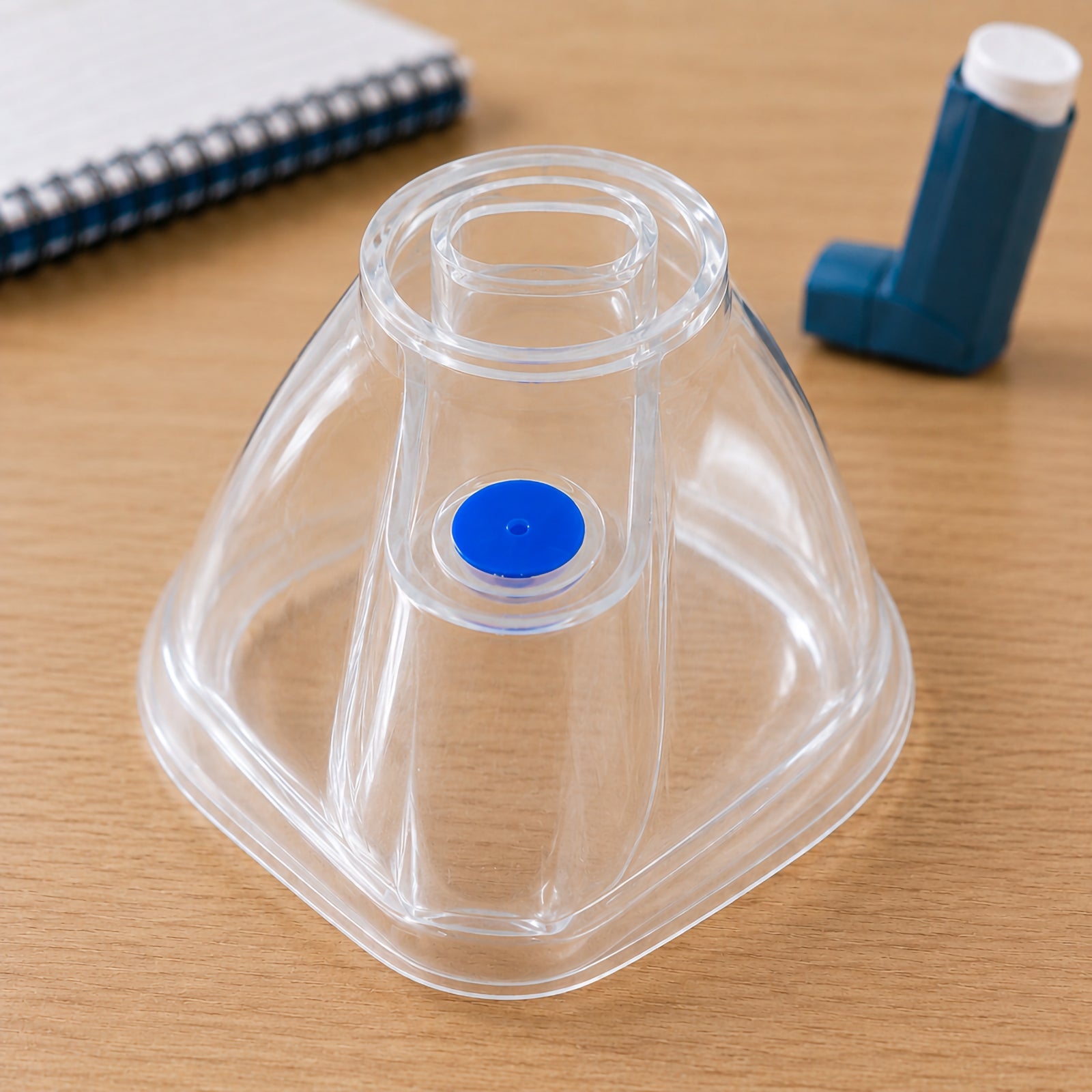 AeroLearn Child Training Mask displayed upright on a timber training desk with a placebo inhaler visible in the background. The image clearly shows the transparent contoured body, circular spacer connection area and blue identifier used to distinguish the child sized model.