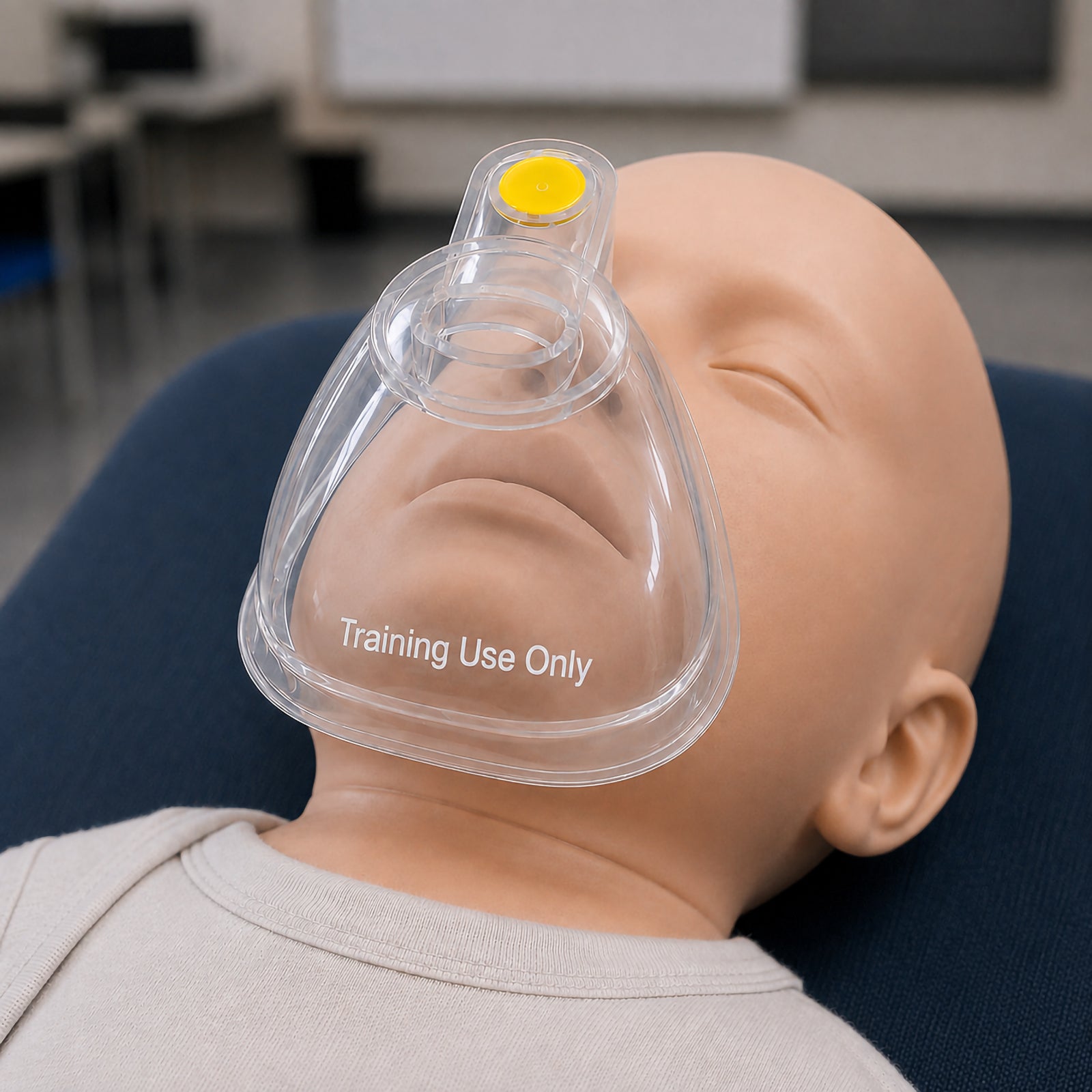 Close view of the AeroLearn Infant Training Mask positioned on an infant manikin. The transparent body provides a clear view of the mouth and surrounding facial area, while the yellow identifier and Training Use Only marking distinguish the equipment as an infant sized respiratory training aid.