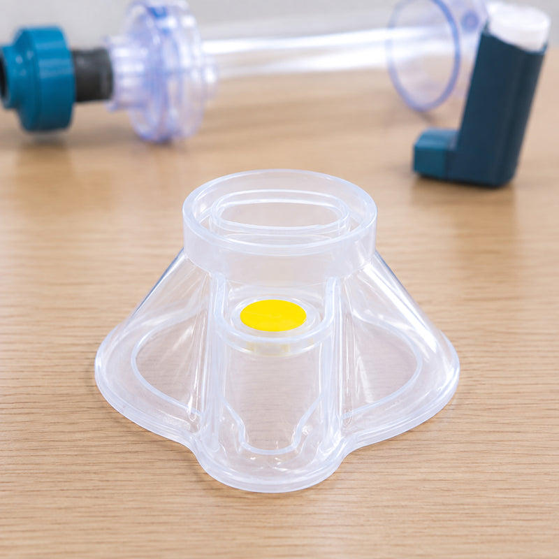 Transparent AeroLearn Infant Training Mask displayed upright on a timber training desk. The compact contoured design, circular connection opening and yellow identifier are clearly visible, with spacer and inhaler training equipment softly shown in the background to provide respiratory education context.