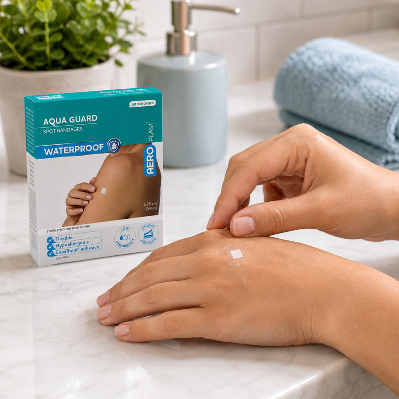 AeroPlast AquaGuard Waterproof Spot Dressings box beside a hand with a transparent circular dressing applied over a small wound pad. The bathroom setting with towels and soap dispenser reinforces the waterproof design and suitability for protecting minor wounds during everyday washing and showering.