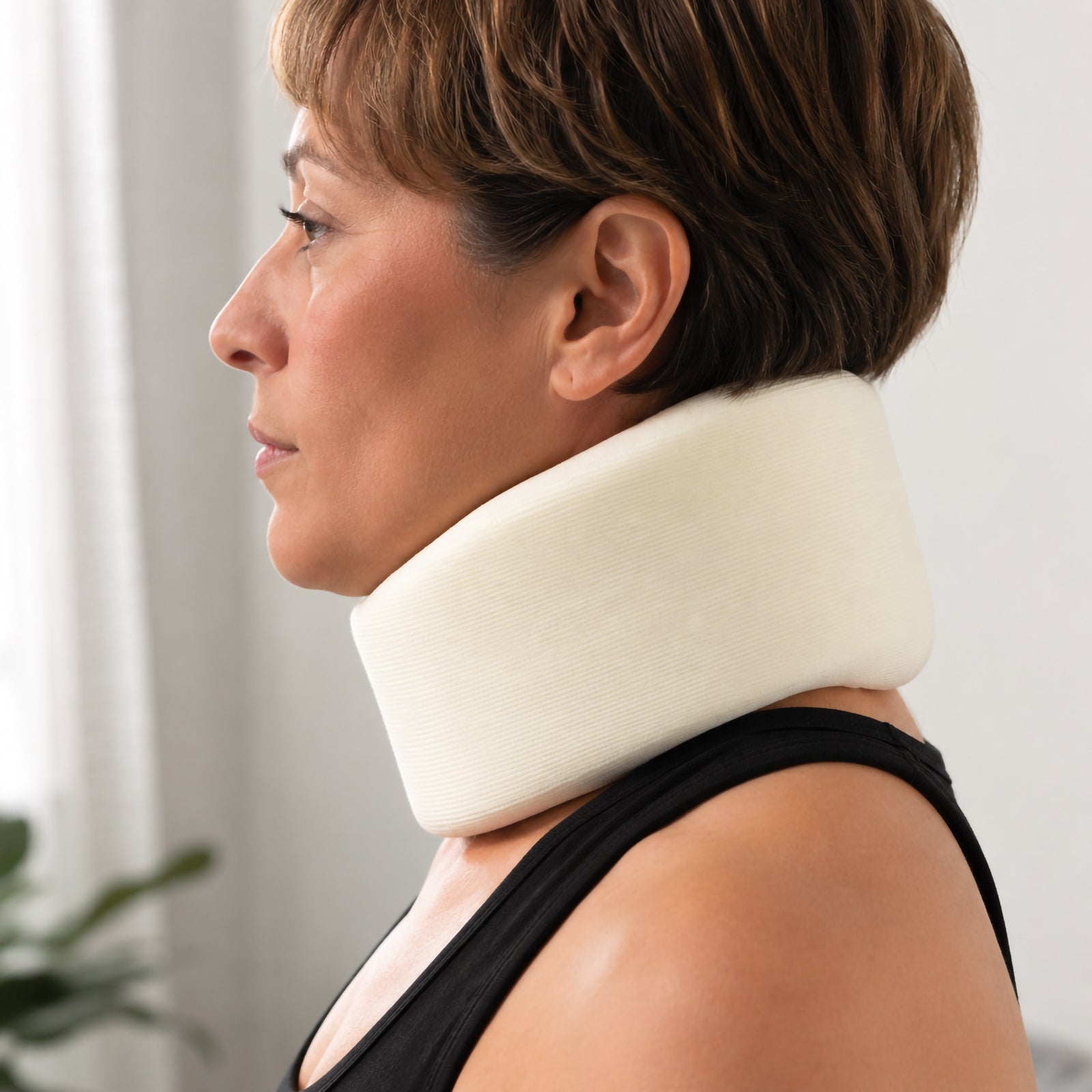 Side profile lifestyle view of a woman wearing the AeroSupplies Foam Cervical Collar Medium, showing the contoured cream foam supporting the chin and neck in a bright home environment.